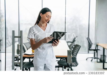 Front view. Standing with notepad in hands. Female doctor in white coat is indoors Front view. Standing with notepad in hands. Female doctor in white coat is indoors 130183397
