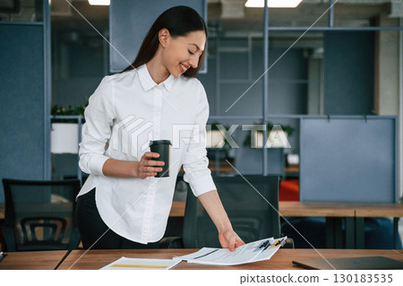 Holding drink in hand. Businesswoman is indoors in the office Holding drink in hand. Businesswoman is indoors in the office 130183535
