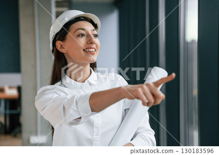 Pointing by finger to the side. Female architect in hard hat and in white clothes is indoors 130183596