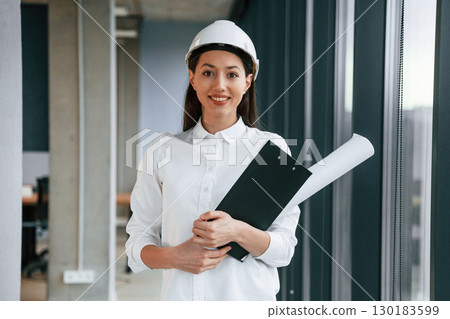 Plan and notepad in hands. Female architect in hard hat and in white clothes is indoors Plan and notepad in hands. Female architect in hard hat and in white clothes is indoors 130183599