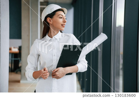 Plan and notepad in hands. Female architect in hard hat and in white clothes is indoors 130183600