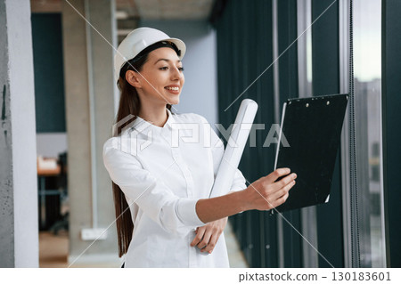 Plan and notepad in hands. Female architect in hard hat and in white clothes is indoors Plan and notepad in hands. Female architect in hard hat and in white clothes is indoors 130183601