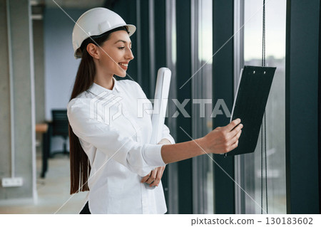 Plan and notepad in hands. Female architect in hard hat and in white clothes is indoors Plan and notepad in hands. Female architect in hard hat and in white clothes is indoors 130183602