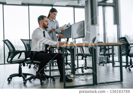 Woman is standing and holding documents. Two employees are working in the office together Woman is standing and holding documents. Two employees are working in the office together 130183789