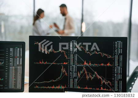 Standing behind the monitors with graphs on them. Two employees are working in the office together Standing behind the monitors with graphs on them. Two employees are working in the office together 130183832