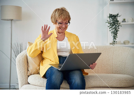 Online call by laptop. Showing hello gesture. Senior elderly woman is at home in the living room 130183985