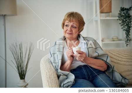 Cup with drink, sitting on a comfortable sofa. Senior elderly woman is at home in the living room 130184005