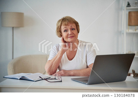 Silver colored laptop. Sitting by the table. Senior elderly woman is at home in the living room Silver colored laptop. Sitting by the table. Senior elderly woman is at home in the living room 130184078