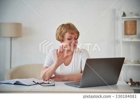 Online call by laptop. Showing hello gesture. Senior elderly woman is at home in the living room 130184081