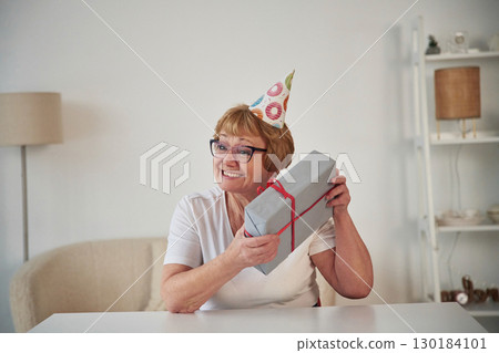 Celebrating birthday. With gift in the present box. Senior elderly woman is at home in the living room Celebrating birthday. With gift in the present box. Senior elderly woman is at home in the living room 130184101