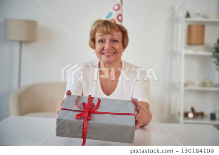 Smiling, celebrating birthday, holding gift box. Senior elderly woman is at home in the living room Smiling, celebrating birthday, holding gift box. Senior elderly woman is at home in the living room 130184109