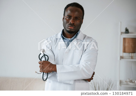 Standing and holding stethoscope. Black man as doctor in the clinic office Standing and holding stethoscope. Black man as doctor in the clinic office 130184971