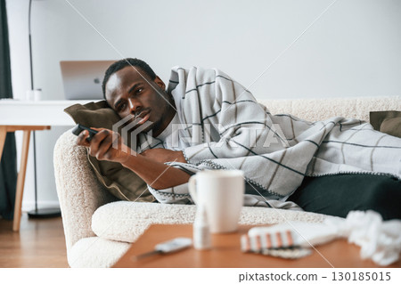 Holding TV remote controller. Lying down. Sick black man is at home Holding TV remote controller. Lying down. Sick black man is at home 130185015
