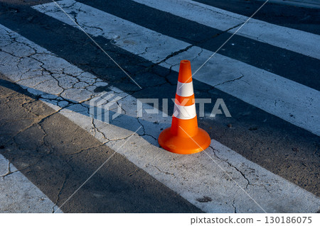 An orange traffic cone sits on cracked pavement at a busy crosswalk under bright sunlight in an urban setting An orange traffic cone sits on cracked pavement at a busy crosswalk under bright sunlight in an urban setting 130186075