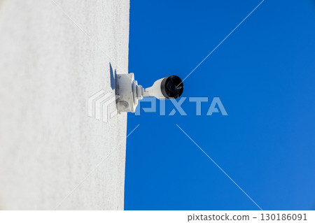 A security camera is installed on a white structure, angled toward a vibrant blue sky, ensuring a watchful eye on its surroundings A security camera is installed on a white structure, angled toward a vibrant blue sky, ensuring a watchful eye on its surroundings 130186091