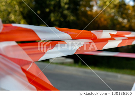 Bright orange and white barrier tape stretches across a path in a park, signaling a restricted area under vibrant trees during midday Bright orange and white barrier tape stretches across a path in a park, signaling a restricted area under vibrant trees during midday 130186100