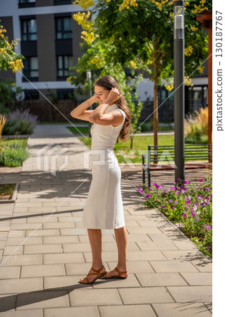 Young woman wearing a white midi dress walking outdoors in Old Money style. Concept of timeless fashion, graceful femininity and understated elegance in modern lifestyle. 130187767