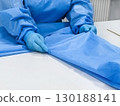 A person in a blue medical gown and gloves is folding a blue medical sheet on a white table.  130188141