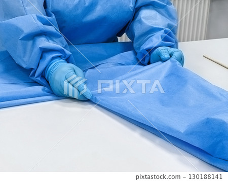 A person in a blue medical gown and gloves is folding a blue medical sheet on a white table.  130188141