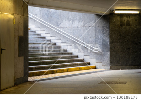 Modern concrete pedestrian staircase inside an urban underpass, with white handrail and tactile paving, leading to a bright exit, depicting urban mobility and safe passage 130188173
