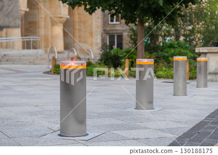A row of modern retractable security bollards with red indicator lights standing on a paved urban square for public access control and safety 130188175