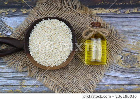 Glass bottle of sesame oil and raw sesame seeds in wooden spoon or bowl on rustic table. Uncooked sesame background concept with copy space Glass bottle of sesame oil and raw sesame seeds in wooden spoon or bowl on rustic table. Uncooked sesame background concept with copy space 130190036