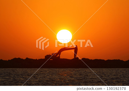An excavator silhouetted on rocks for marina construction at sunset. With the sun shining brightly. An excavator silhouetted on rocks for marina construction at sunset. With the sun shining brightly. 130192378