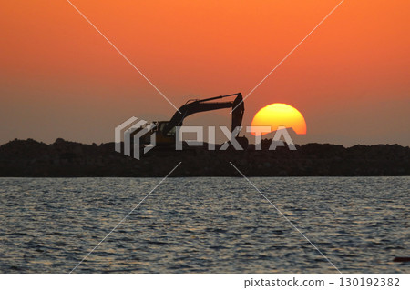 An excavator silhouetted on rocks for marina construction at sunset. With the sun shining brightly. An excavator silhouetted on rocks for marina construction at sunset. With the sun shining brightly. 130192382