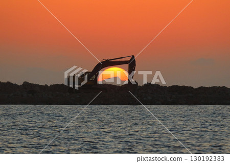 An excavator silhouetted on rocks for marina construction at sunset. With the sun shining brightly. 130192383
