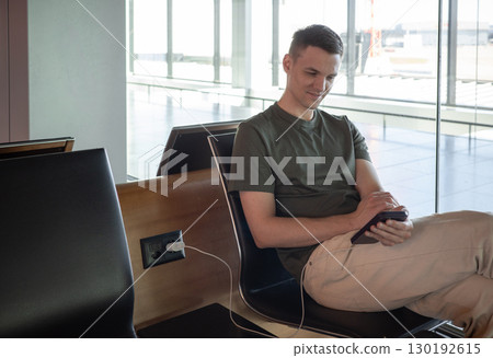 Young man charging phone at airport, using smartphone while waiting for flight. Comfortable travel concept in modern terminal. 130192615