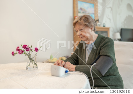 Senior woman measures her blood pressure while sitting at home in comfortable environment. Concept of health, wellness, self-care, medical monitoring, and senior independence. Healthcare for seniors. 130192618