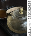Close up of textured glass teapot with golden lid and rope handle on a wooden tray. Concept of tea ceremony, warmth, and cozy atmosphere. 130192626