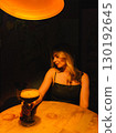 Young woman sitting at round wooden table in dimly lit bar, holding large stemmed glass containing espresso martini cocktail, illuminated by orange lamp above. Woman holding espresso martini cocktail 130192645