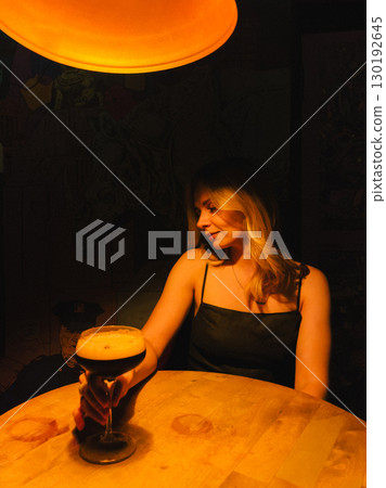 Young woman sitting at round wooden table in dimly lit bar, holding large stemmed glass containing espresso martini cocktail, illuminated by orange lamp above. Woman holding espresso martini cocktail 130192645