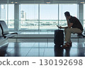 Young man sitting at airport gate waiting for a flight, looking out the window at airplanes with smartphone and suitcase. Modern air travel concept. 130192698