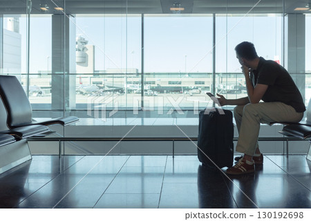 Young man sitting at airport gate waiting for a flight, looking out the window at airplanes with smartphone and suitcase. Modern air travel concept. 130192698