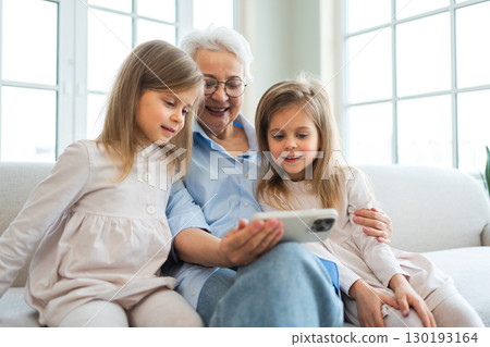 Happy family at home. Two little girls sisters twins grandmother enjoying time together watching video on phone. Good time at home. Grandma granddaughters child kids together. Family generations Happy family at home. Two little girls sisters twins grandmother enjoying time together watching video on phone. Good time at home. Grandma granddaughters child kids together. Family generations 130193164
