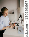 Young woman assembling plastic model kit at home, carefully following instructions on booklet on white table with pieces scattered around and lamp in the background. Woman constructing plastic model 130193667