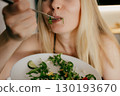 Blond woman savoring fresh salad with fork from white plate, relishing nutritious meal in her kitchen, surrounded by vibrant vegetables and wholesome ingredients. Blond woman enjoying fresh salad 130193670