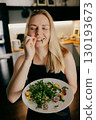 Happy woman with closed eyes chewing on leaf of arugula while holding plate of fresh salad in modern kitchen, enjoying healthy meal. Woman savoring arugula leaf, experiencing wholesome dining moment 130193673