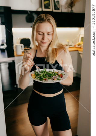Blond woman wearing sportswear is holding plate of fresh mixed salad and eating it with closed eyes and delightful expression in her modern kitchen. Sporty woman enjoying mixed salad. Healthy life 130193677