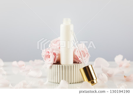 White dropper bottle with serum on a marble podium on a white table with tender pink roses and rose petals. Natural beauty product based on rose flowers. Soft focus style 130194190