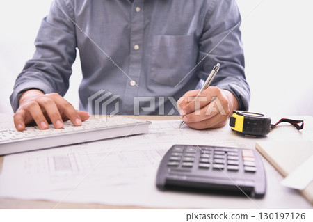 A professional working on project plans with calculator, keyboard and measuring tape in a modern office setting. A professional working on project plans with calculator, keyboard and measuring tape in a modern office setting. 130197126