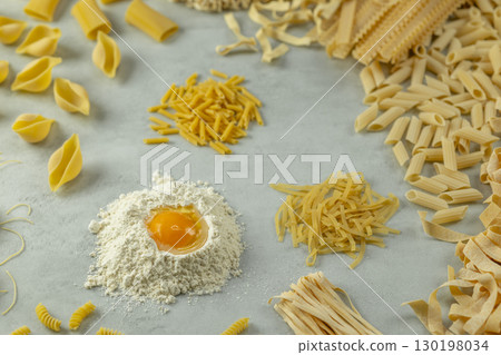 Variety of uncooked pasta with flour and egg yolk forming a circle on a gray background 130198034