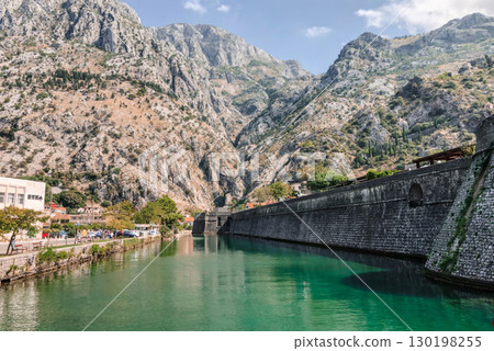 A picturesque view of the ancient city walls of Kotor, Montenegro, rising majestically alongside a tranquil canal with emerald green waters. A picturesque view of the ancient city walls of Kotor, Montenegro, rising majestically alongside a tranquil canal with emerald green waters. 130198255