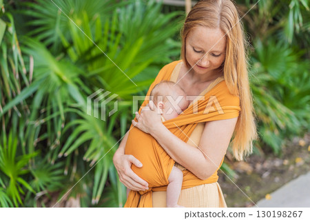 Mother holding her little baby in a yellow sling in the park. Warm and loving family moment. Babywearing, parenting, and mother-child bonding concept 130198267