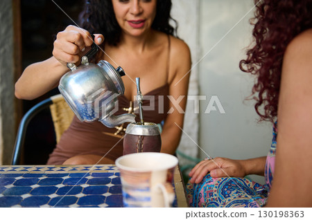 Woman Preparing Traditional Beverage Mate with Kettle and Friends at Outdoor Gathering Woman Preparing Traditional Beverage Mate with Kettle and Friends at Outdoor Gathering 130198363