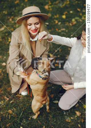 Woman, little girl and dog playing in the autumn park. Brown french bulldog with female owners spend a day at the park playing and having fun. Woman wearing beige coat and a hat. 130199477