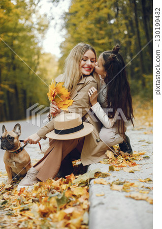 Woman, little girl and dog playing in the autumn park. Brown french bulldog with female owners spend a day at the park playing and having fun. Woman wearing beige coat and a hat. 130199482