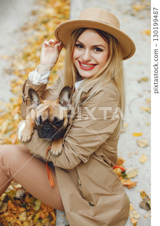 Woman and dog play and have fun in the autumn park. Brown french bulldog with female owner spend a day at the park playing and having fun. Woman wearing beige coat and a hat. 130199487